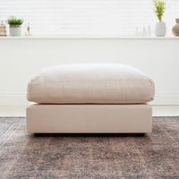 A Hampshire Modular Footstool in Beige with a plush duck feather cushion sits on a patterned rug by a bright window.