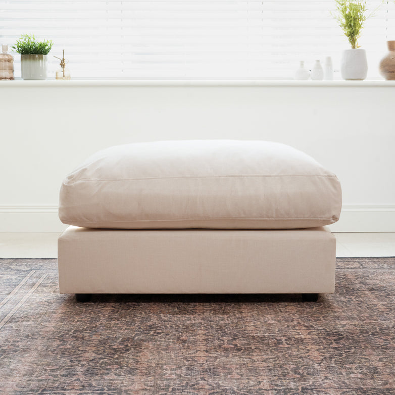 A Hampshire Modular Footstool in Beige with a plush duck feather cushion sits on a patterned rug by a bright window.