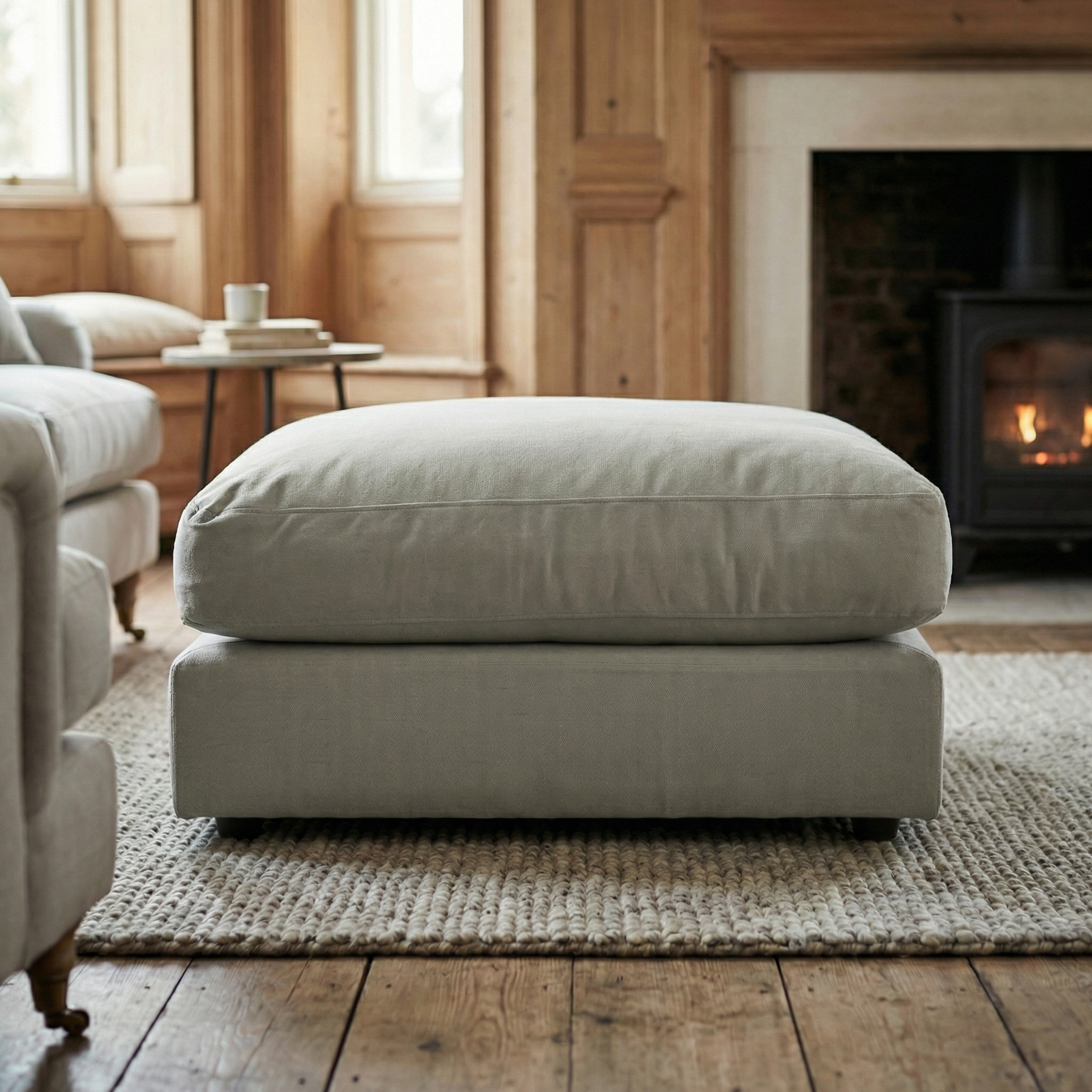 Large Hampshire Modular Footstool in Classic Grey on a textured rug in a cozy living room with wood paneling, fireplace, and plush duck feather cushion. Handmade in England for timeless charm.