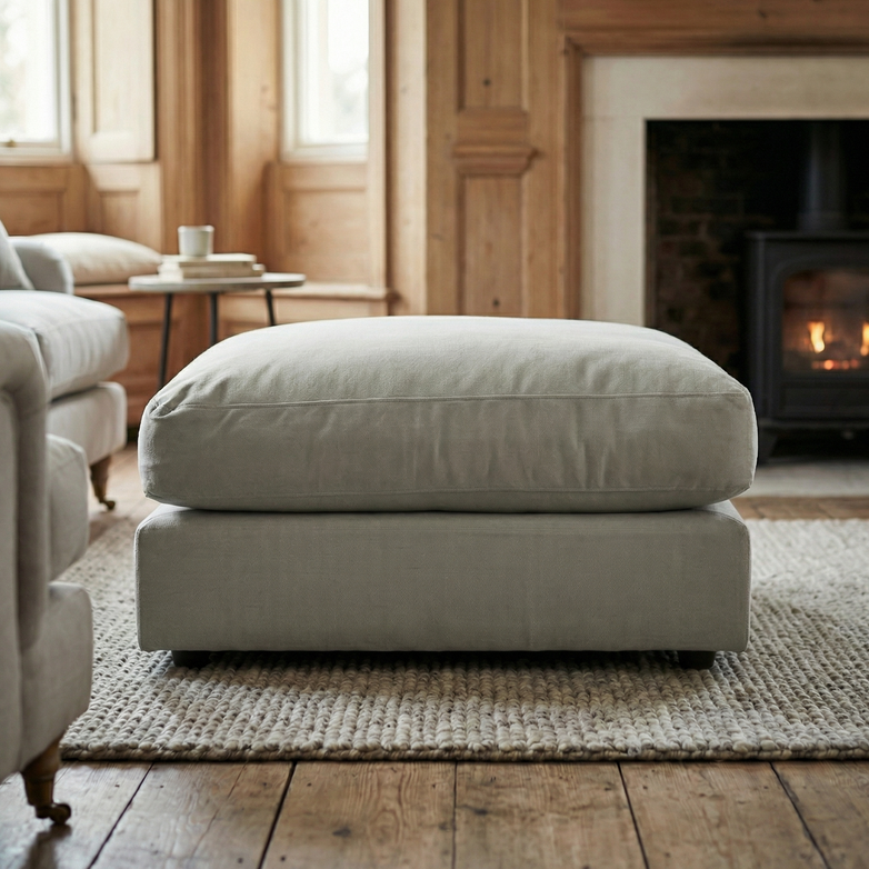 Large Hampshire Modular Footstool in Classic Grey on a textured rug in a cozy living room with wood paneling, fireplace, and plush duck feather cushion. Handmade in England for timeless charm.