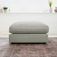 Classic grey Hampshire Modular Footstool with a plush duck feather cushion sits on a patterned rug, framed by a bright window and stylish decor in the background.