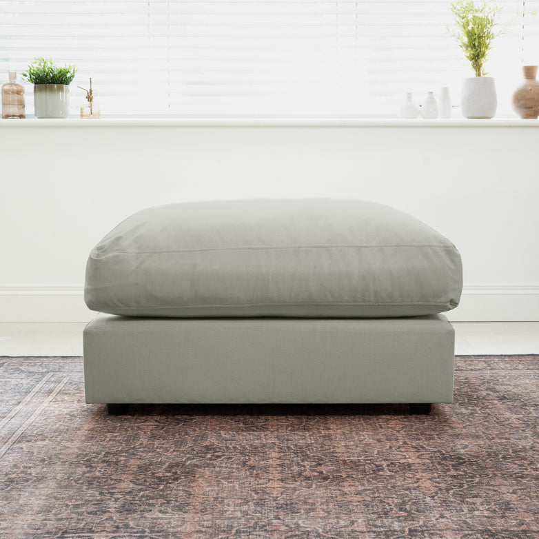 Classic grey Hampshire Modular Footstool with a plush duck feather cushion sits on a patterned rug, framed by a bright window and stylish decor in the background.