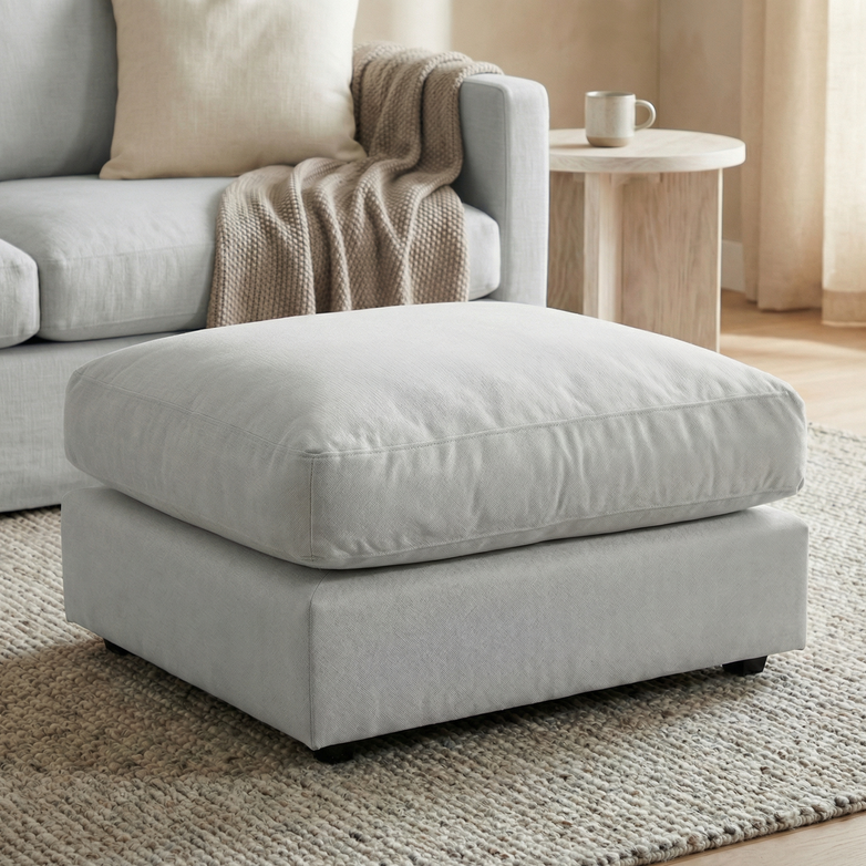 The Hampshire Modular Footstool in Dove Grey, handmade in England with linen-effect fabric, rests on a textured rug beside a blanket-draped sofa and a side table topped with a mug.