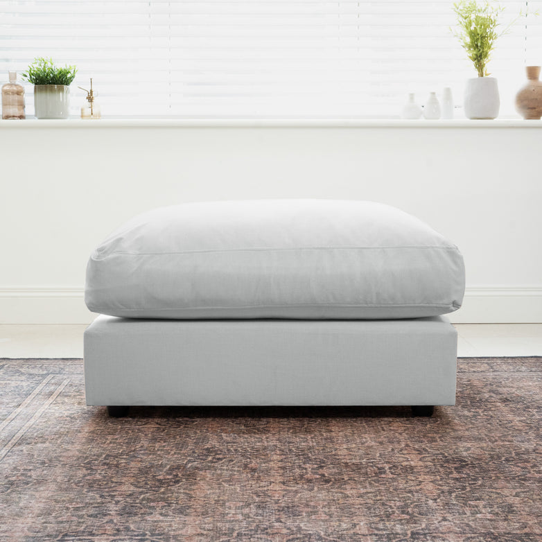 Dove Grey Hampshire Modular Footstool on a rug before a bright window, accented by plants and vases on the sill; handmade in England for a luxurious touch.
