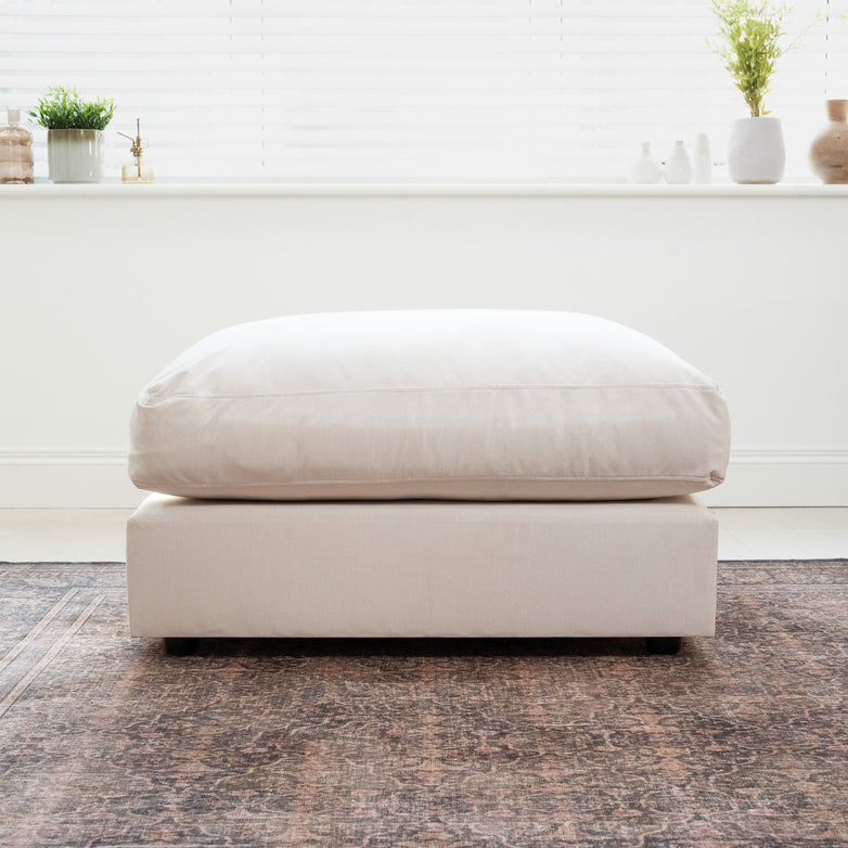 The Hampshire Modular Footstool in Ivory, featuring a plush cushion, sits atop a patterned rug in a bright, modern room filled with plants and stylish decor.