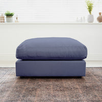 The Hampshire Modular Footstool in Midnight Blue with a duck feather cushion sits on a patterned rug in a bright room, complemented by plants on the windowsill.