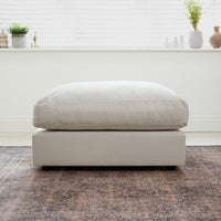 The Hampshire Modular Footstool in Warm Grey rests on a patterned rug in a bright, modern room with plants on the windowsill.