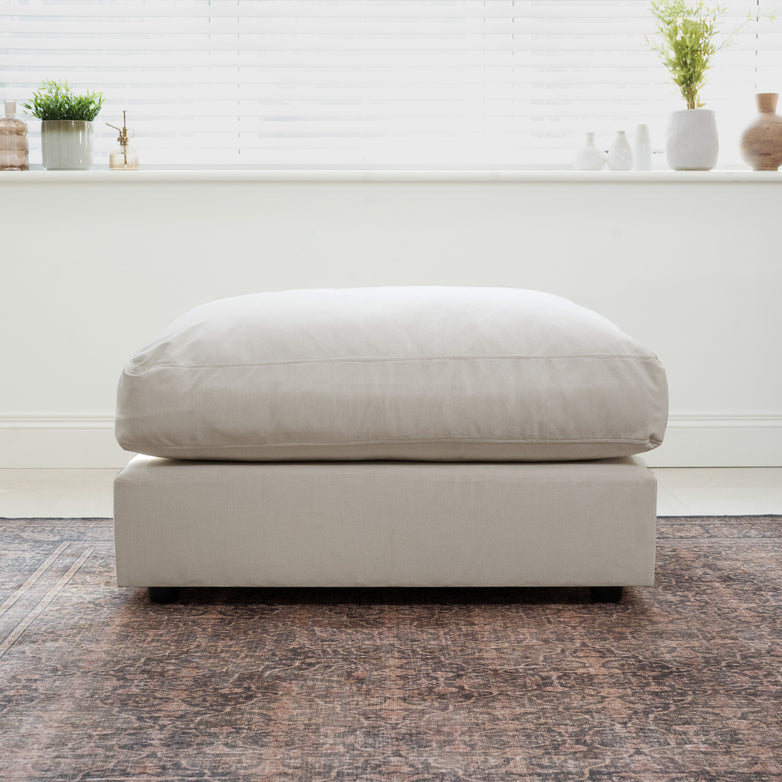 The Hampshire Modular Footstool in Warm Grey rests on a patterned rug in a bright, modern room with plants on the windowsill.