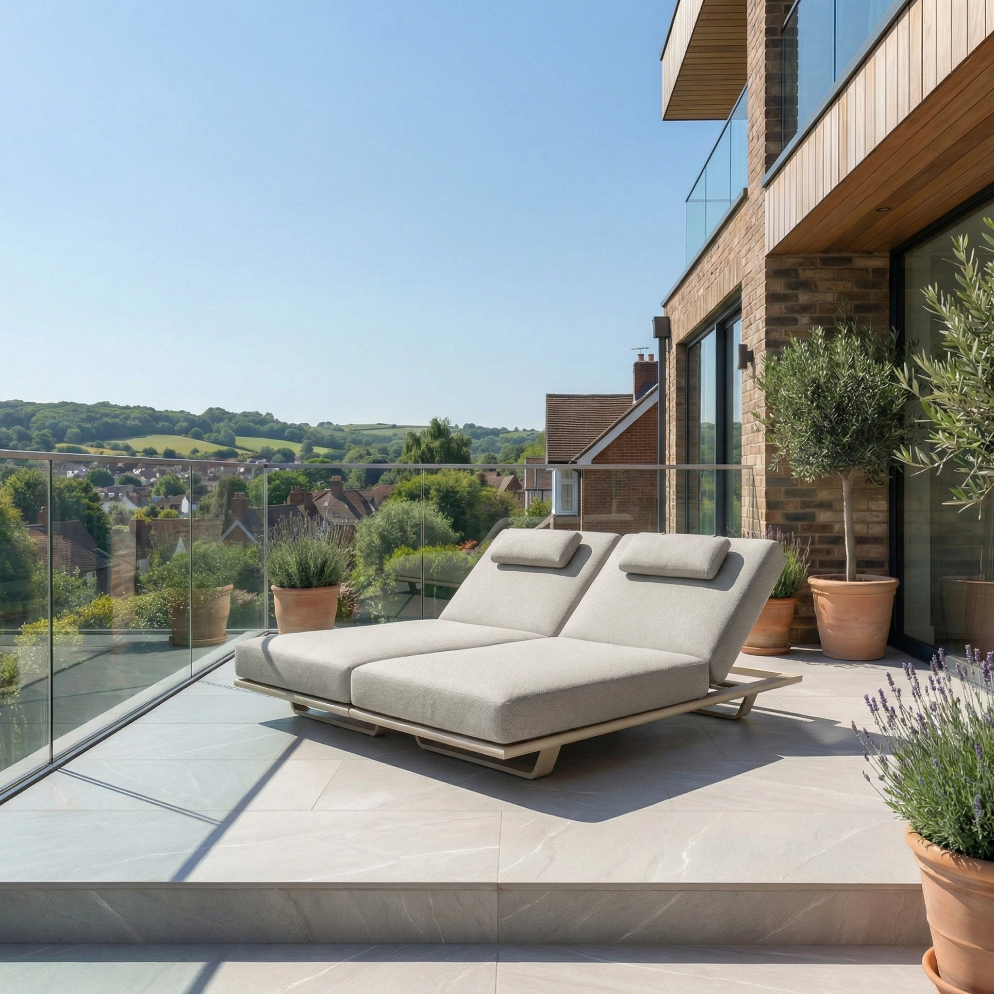 The Hatia Double Sun Lounger in Latte with Olefin cushions sits on a sunny terrace with glass railing, offering scenic countryside views.
