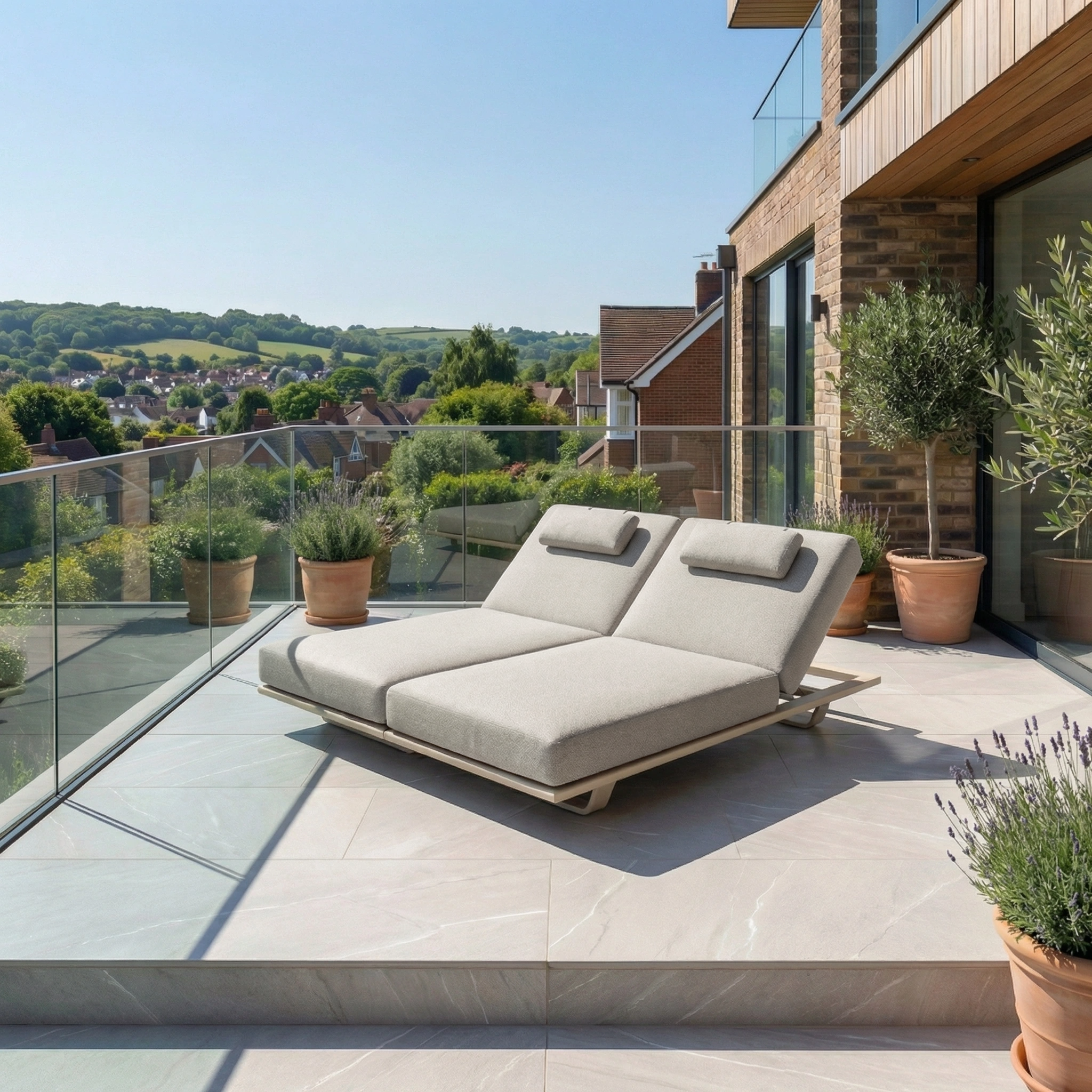 Two Hatia Double Sun Loungers in Latte with Olefin cushions are placed on a sunny terrace with a glass railing, surrounded by potted plants and scenic greenery.