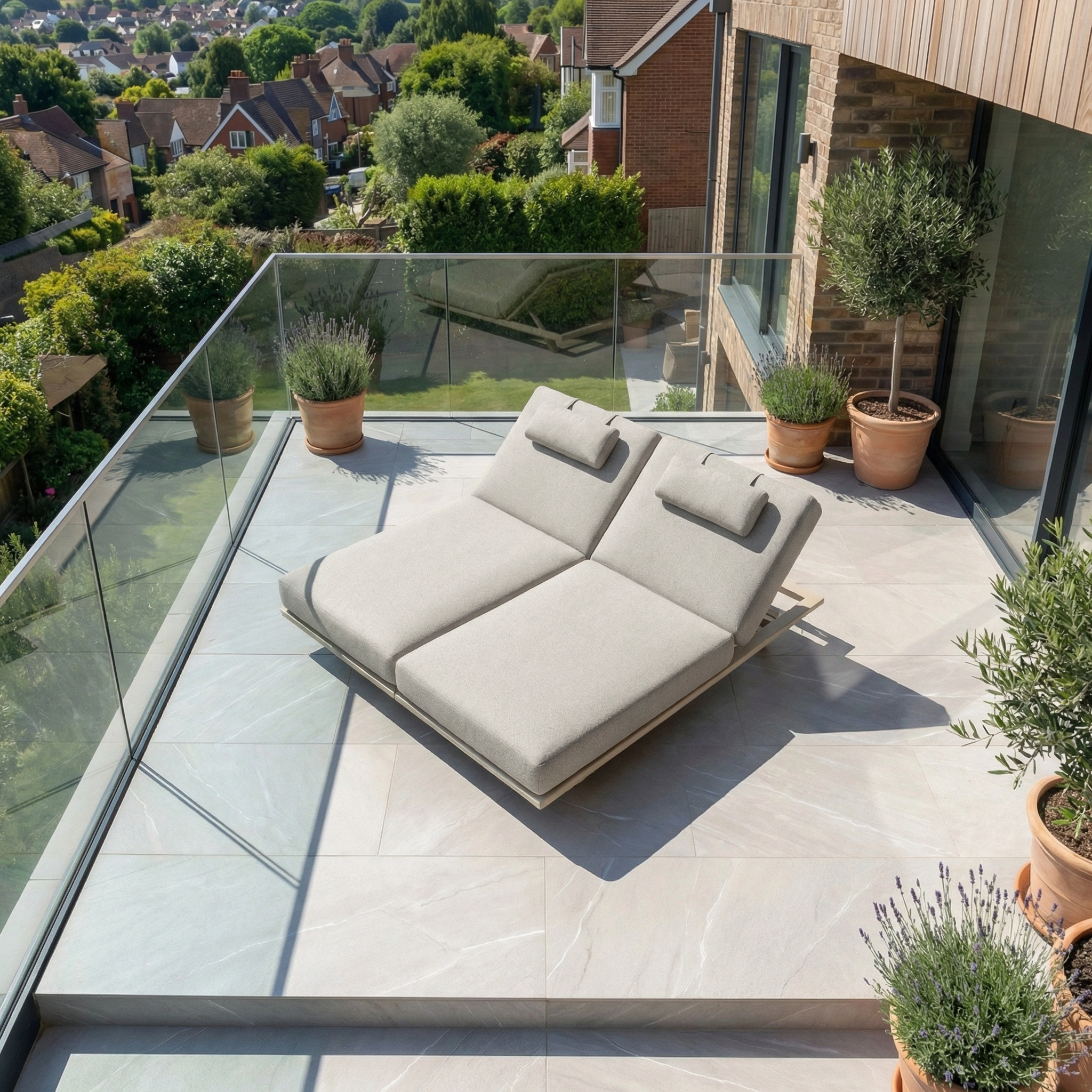 Modern outdoor balcony with the Hatia Double Sun Lounger in Latte featuring plush olefin cushions, potted plants, and views of houses and greenery.