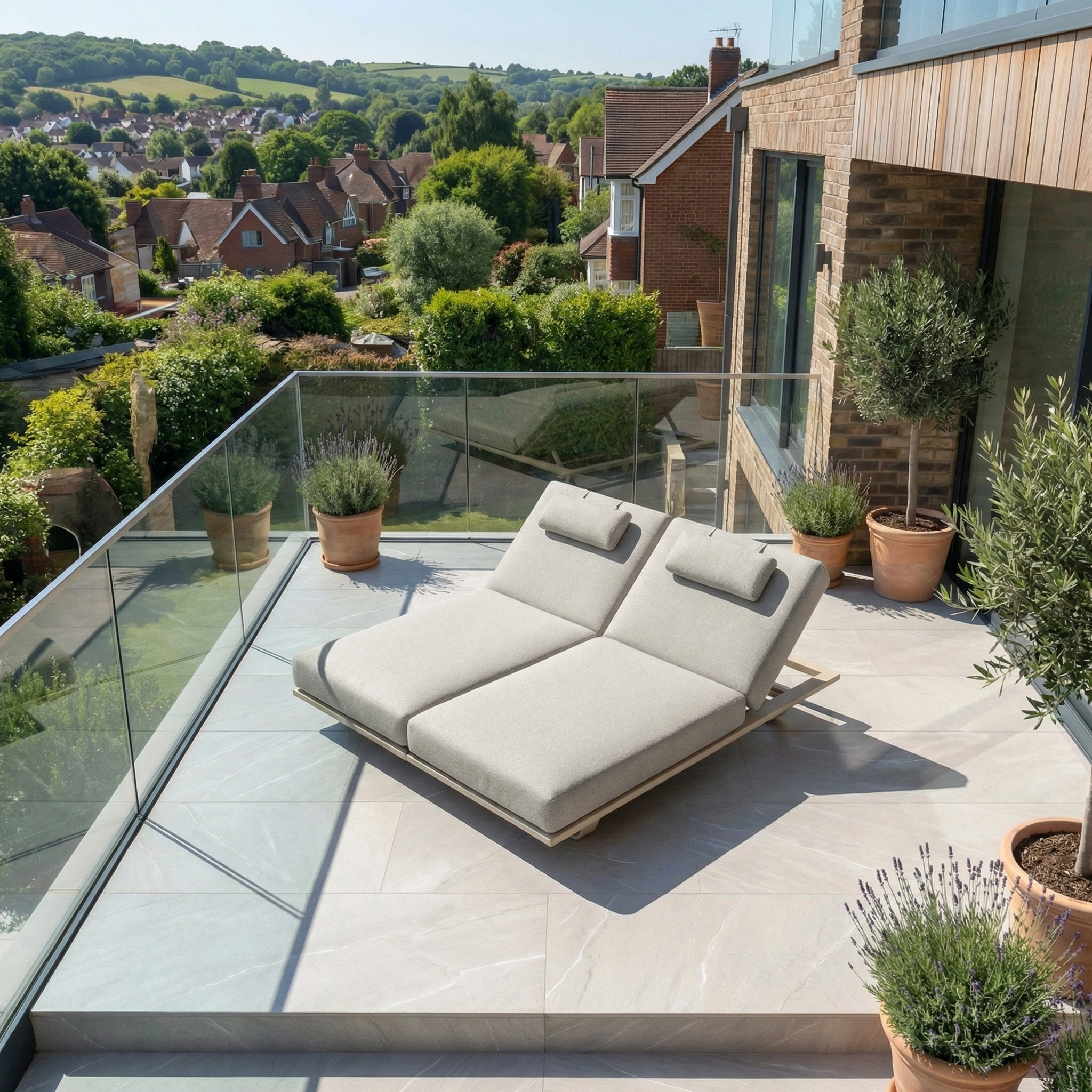 The Hatia Double Sun Lounger in Latte, featuring plush olefin cushions, sits on a modern balcony with potted plants and overlooks a scenic suburban neighborhood.