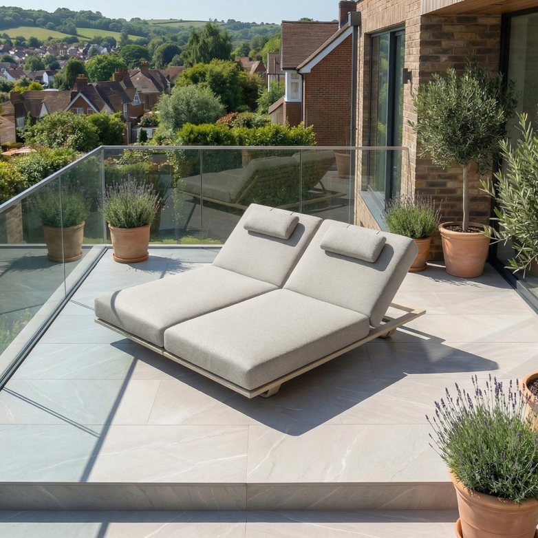 The Hatia Double Sun Lounger in Latte, featuring plush Olefin cushions, sits on a tiled balcony with potted plants, offering scenic views of nearby houses and lush green hills.