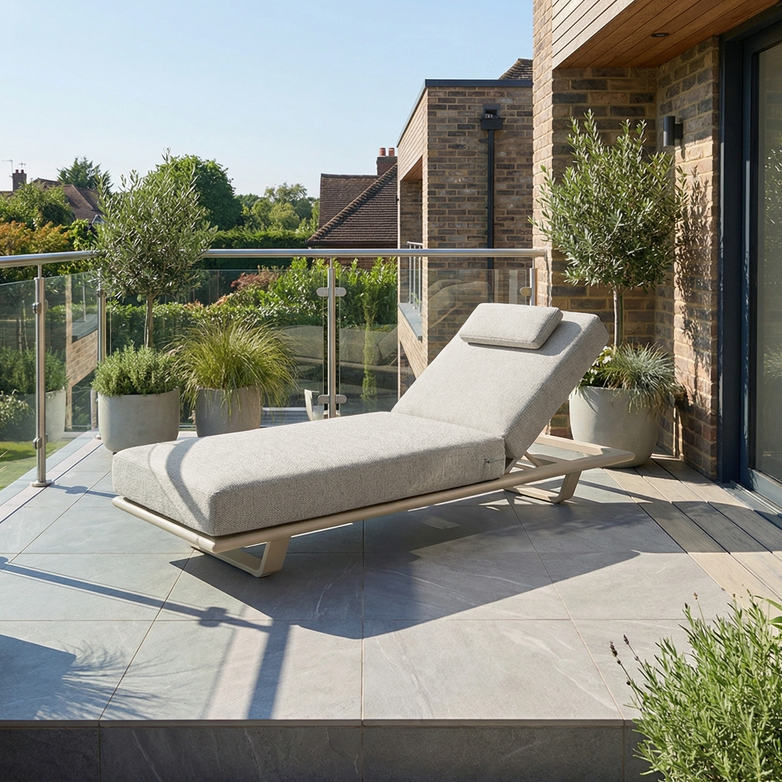 The Hatia Single Sun Lounger in Latte with cushions sits on a tiled balcony amid potted plants and a glass railing. This UV-resistant chair offers stylish, durable outdoor relaxation.