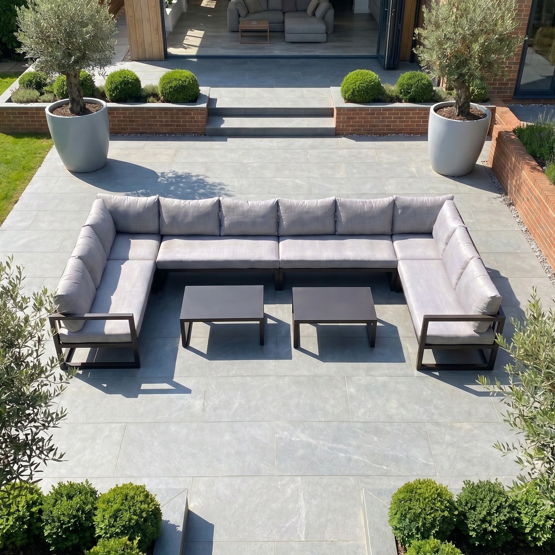 The Havana U-Shape Corner Group Set with Reclining Feature and Coffee Tables in Washed Grey creates a stylish, relaxing outdoor space on your patio, surrounded by plants and brick walls—ideal for unwinding in comfort.