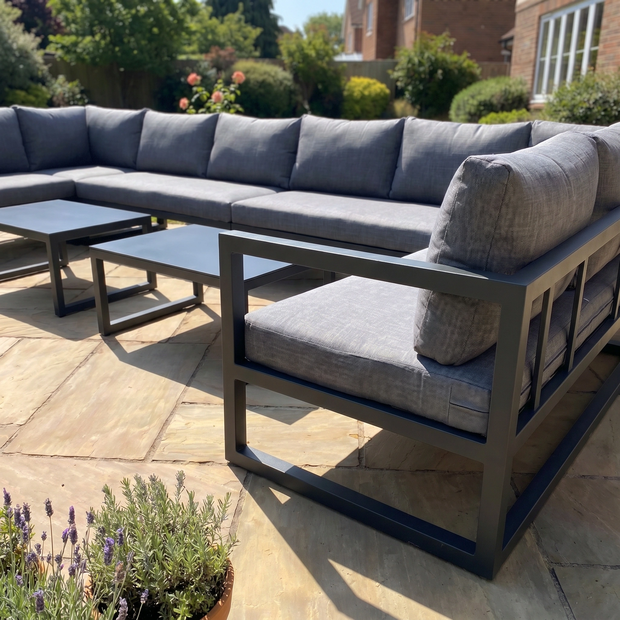 The Havana U-Shape Corner Group Set in charcoal features a reclining function and coffee tables, offering outdoor luxury on any patio and creating an inviting space surrounded by garden plants and flowers.