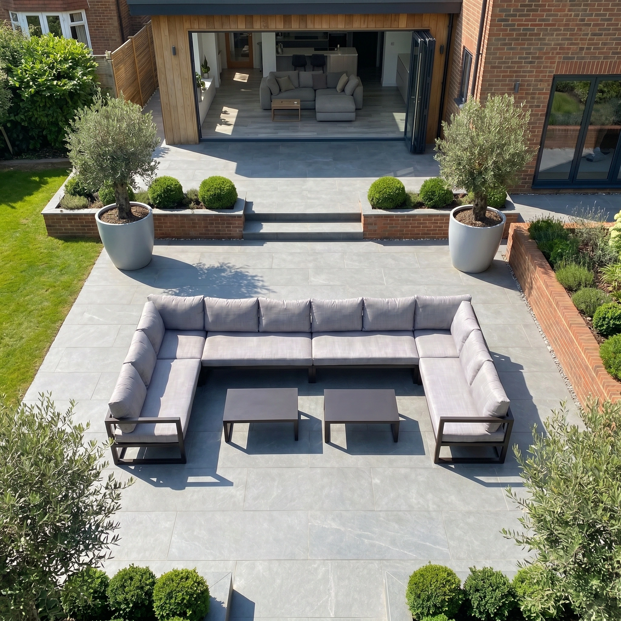 The Havana U-Shape Corner Group Set with Reclining Feature and Coffee Tables in Washed Grey is shown on a grey tiled patio, surrounded by potted olive trees, neat boxwood bushes, and a stylish outdoor coffee table.