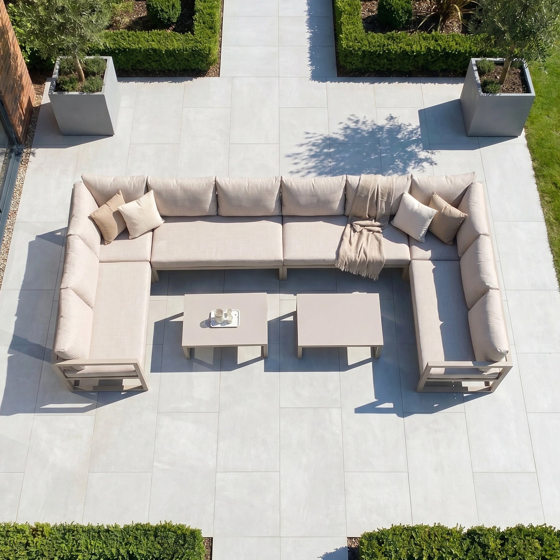 The Havana U-Shape Corner Group Set with Reclining Feature and Coffee Tables in Latte sits on a tiled patio, surrounded by bushes and potted trees, offering style and comfort with plush UV-resistant cushions.