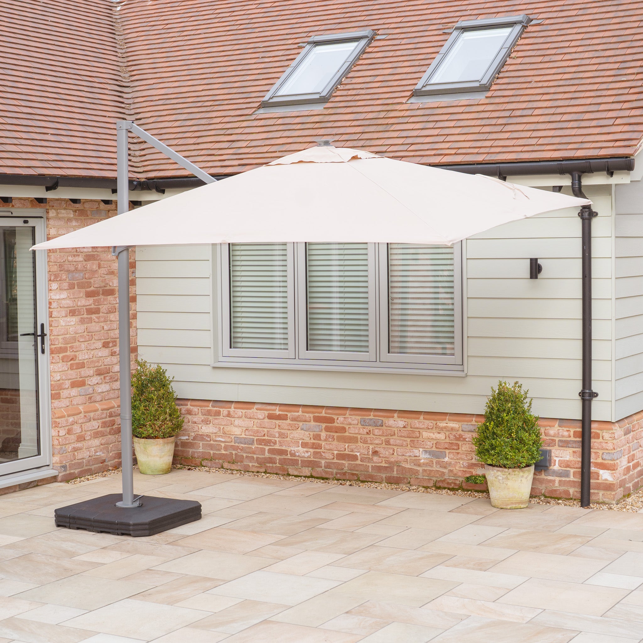 The patio showcases a beige Hera 2.7m Square Cantilever Parasol with LED lighting, UV50+ fabric, two potted plants, and a modern house with skylights and patio doors elegantly in the background.