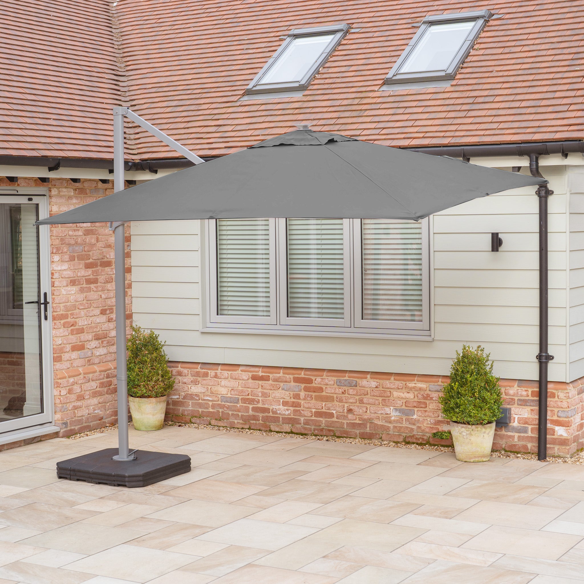 The Hera 2.7m Square Cantilever Parasol with LED Lighting in Charcoal stands open on a stone terrace before a house with brick and siding walls.