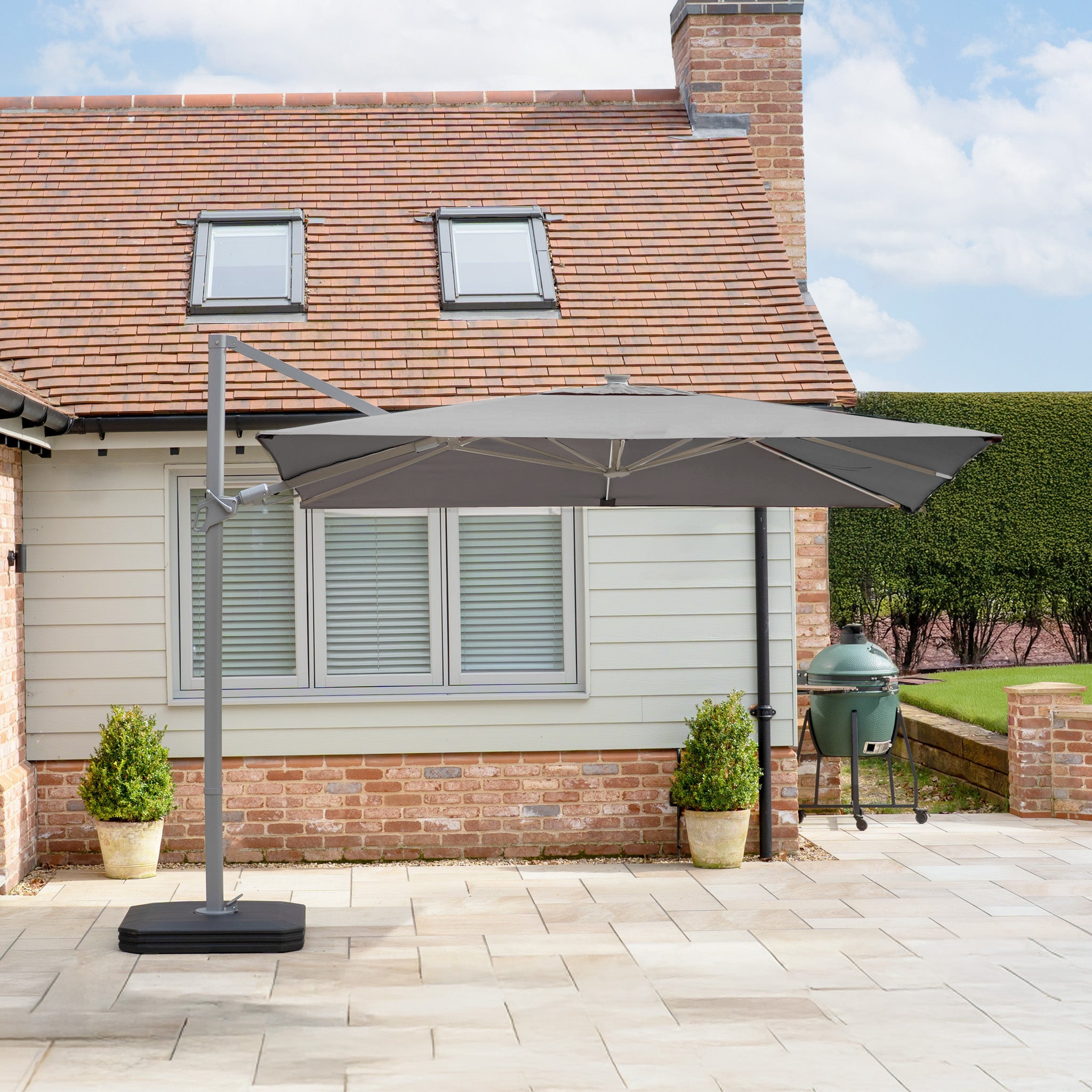 The patio showcases the Hera 3m x 2m Rectangular Cantilever Parasol in Charcoal, featuring LED lighting beside a brick house, complemented by potted plants and a green barbecue grill under the clear sky.