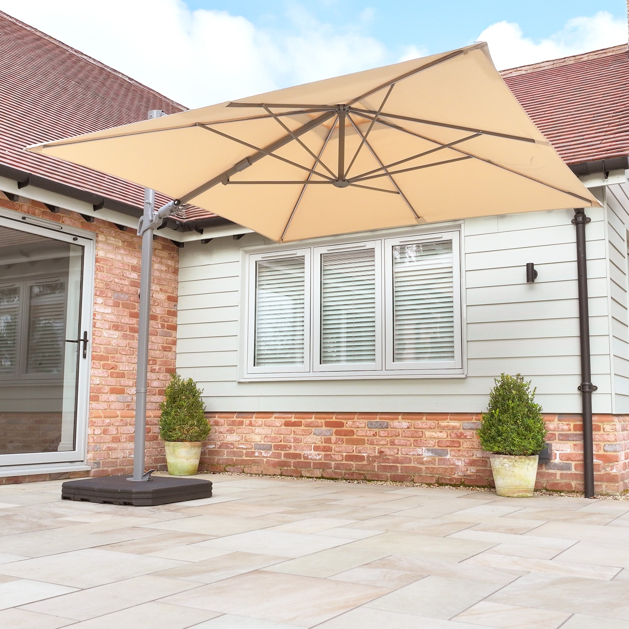 A patio with a Hera 2.7m Square Cantilever Parasol featuring UV50+ fabric and LED lighting in beige, set against a brick and siding house, complemented by two potted plants and a tiled floor.