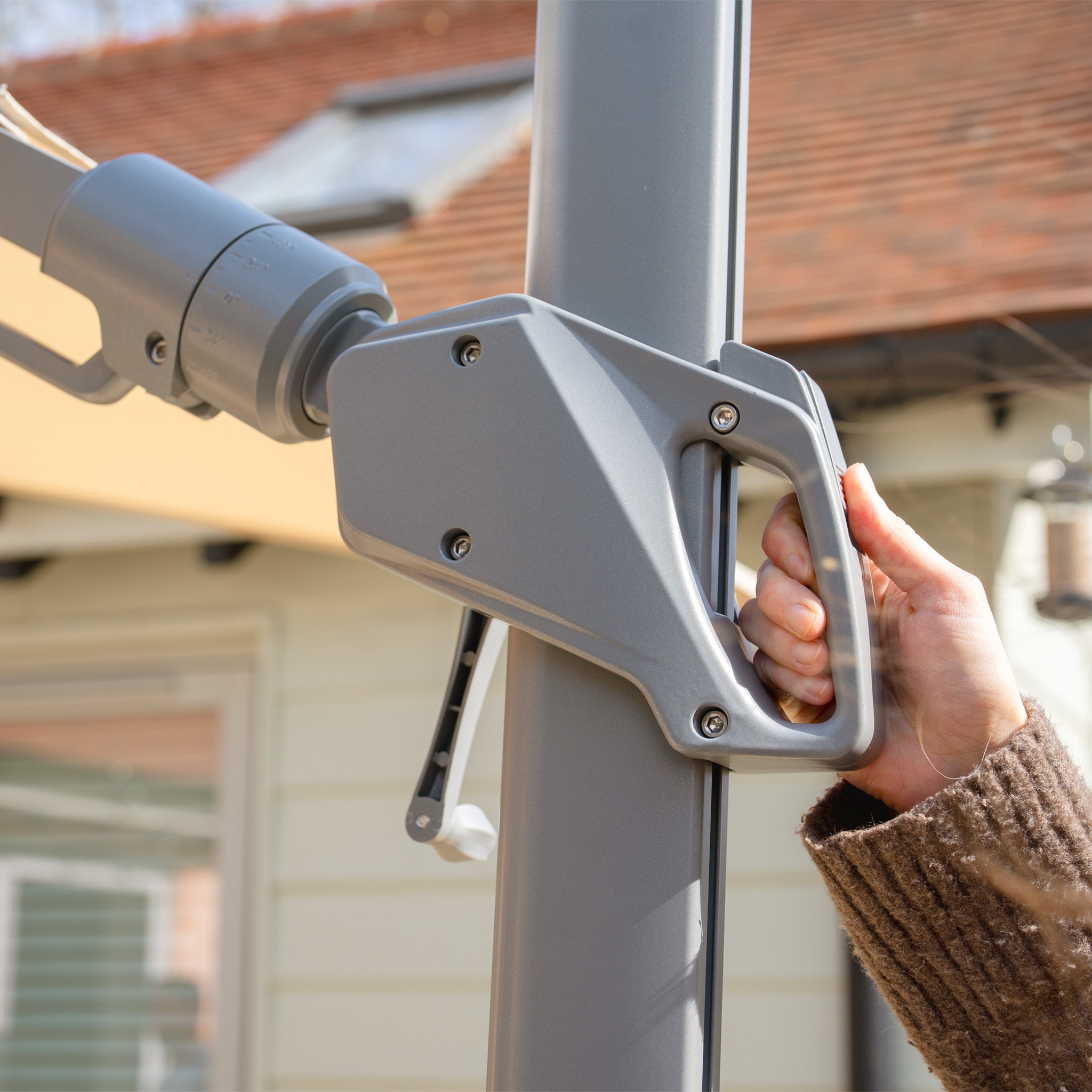 A close-up shows a person's hand operating the crank on a gray pole, highlighting the sleek design of the Hera 2.7m Square Cantilever Parasol with LED Lighting in Beige, set in a backyard setting.