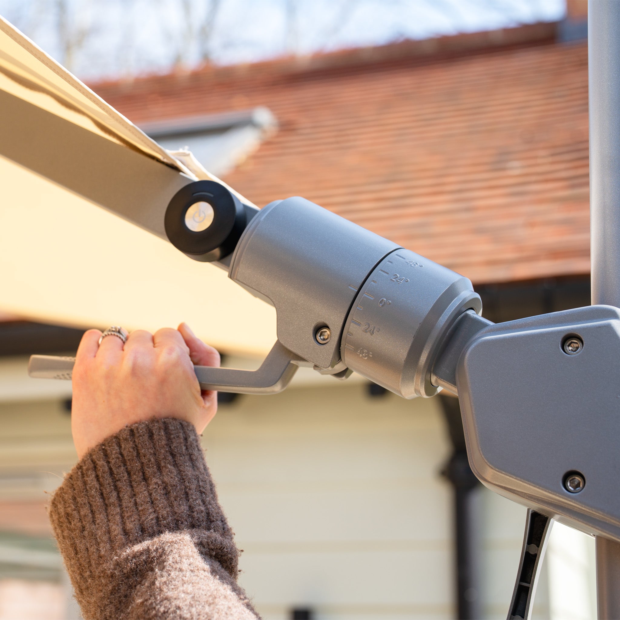 A person adjusts the crank on a beige Hera 2.7m Square Cantilever Parasol with LED Lighting, enjoying UV50+ fabric shade on a sunny day near a house roof.