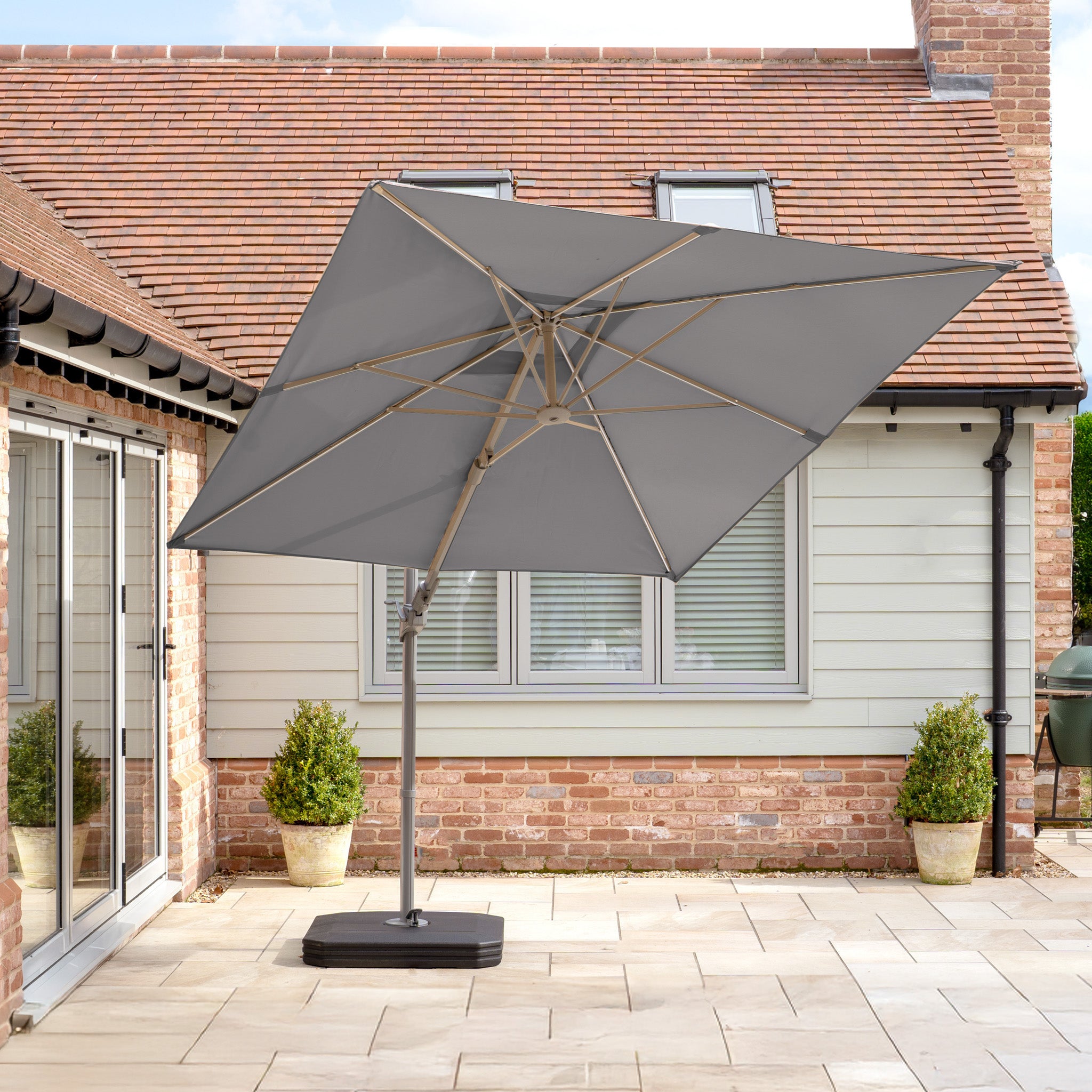 The Hera 3m x 2m Rectangular Cantilever Parasol in Charcoal casts a charming ambiance on a patio with potted plants, providing shade next to the house's brick walls and large windows.