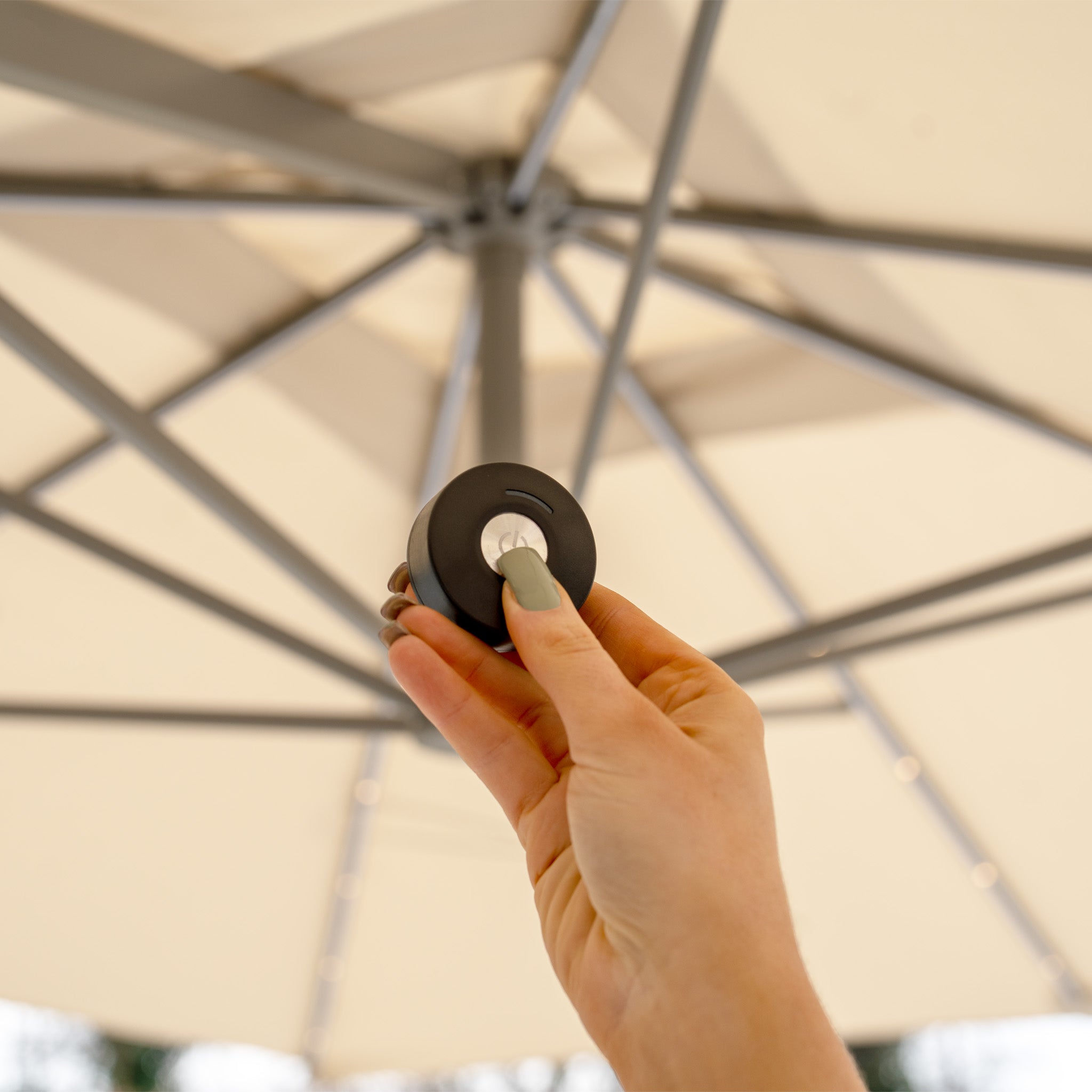 A hand holds a sleek black remote under a beige Hera 2.7m Square Cantilever Parasol, its geometric frame flawlessly enhancing the subtle glow of built-in LED lights.
