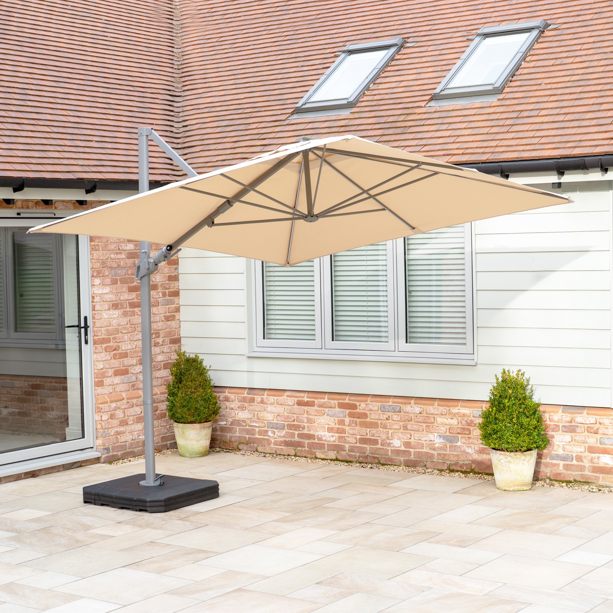 A beige Hera 2.7m Square Cantilever Parasol with LED lighting stands by a brick and siding house with a tiled roof and skylights, providing shade with its UV50+ fabric.
