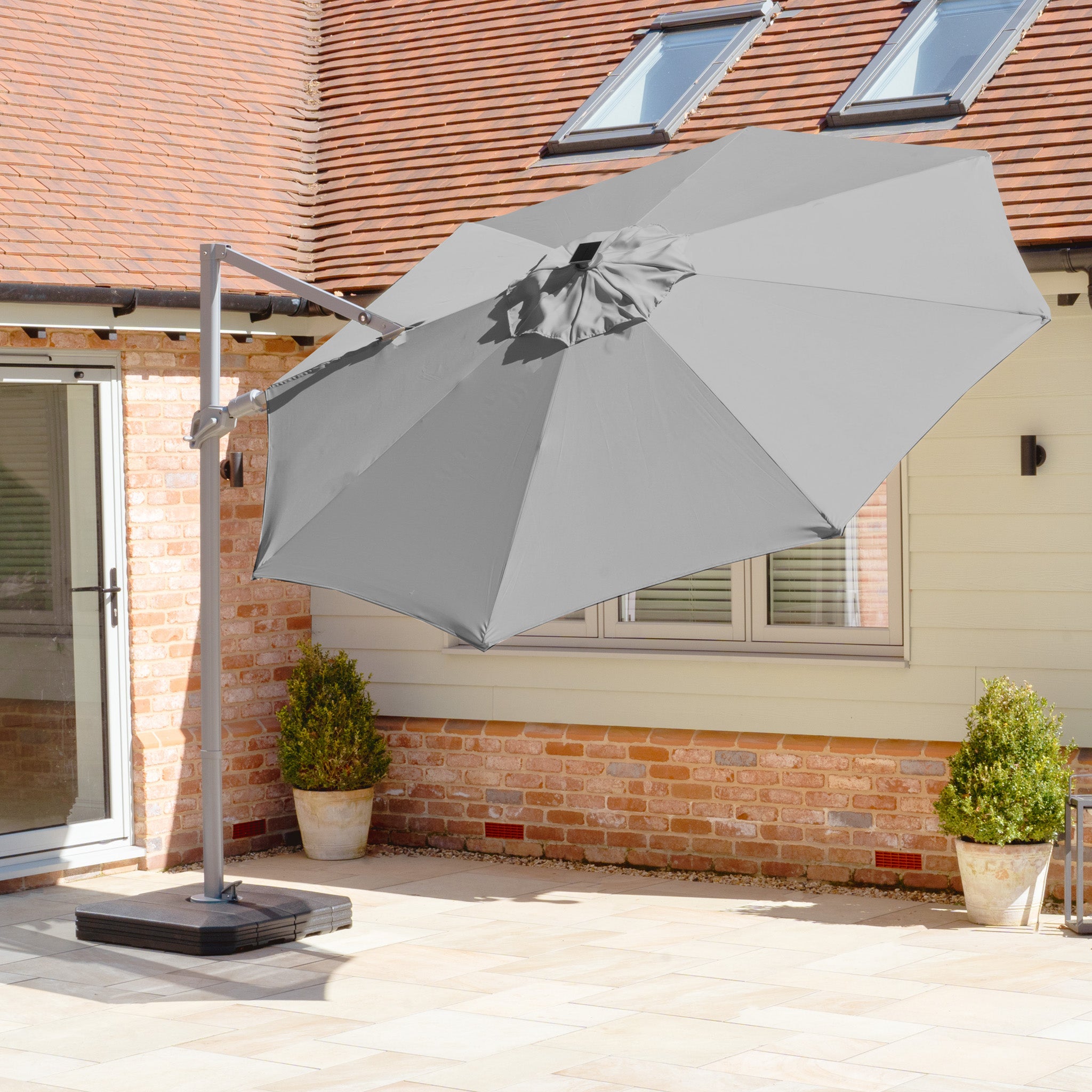 The Hera 3m Round Cantilever Parasol with LED Lighting in Charcoal provides UV50+ sun protection, casting shade across the sunny terrace, while potted plants enhance the brick wall's greenery. Enjoy your outdoor space comfortably all day with this stylish umbrella.