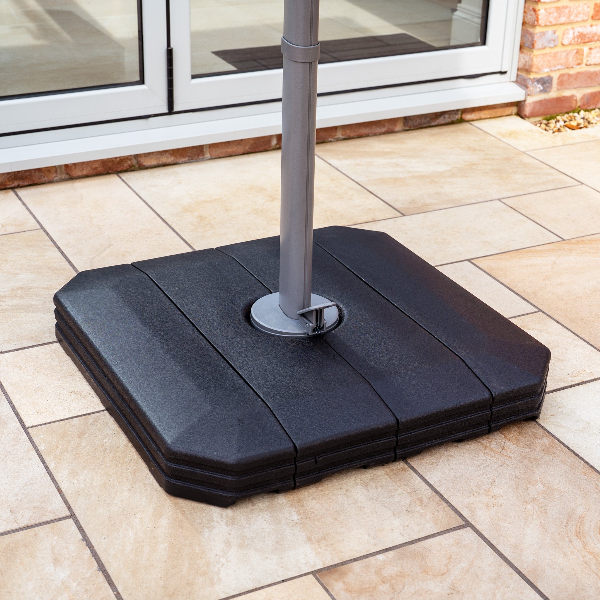 The Hera 3m Round Cantilever Parasol with LED Lighting in Beige is supported by a black square base with metal pole on beige outdoor tiles near glass doors.