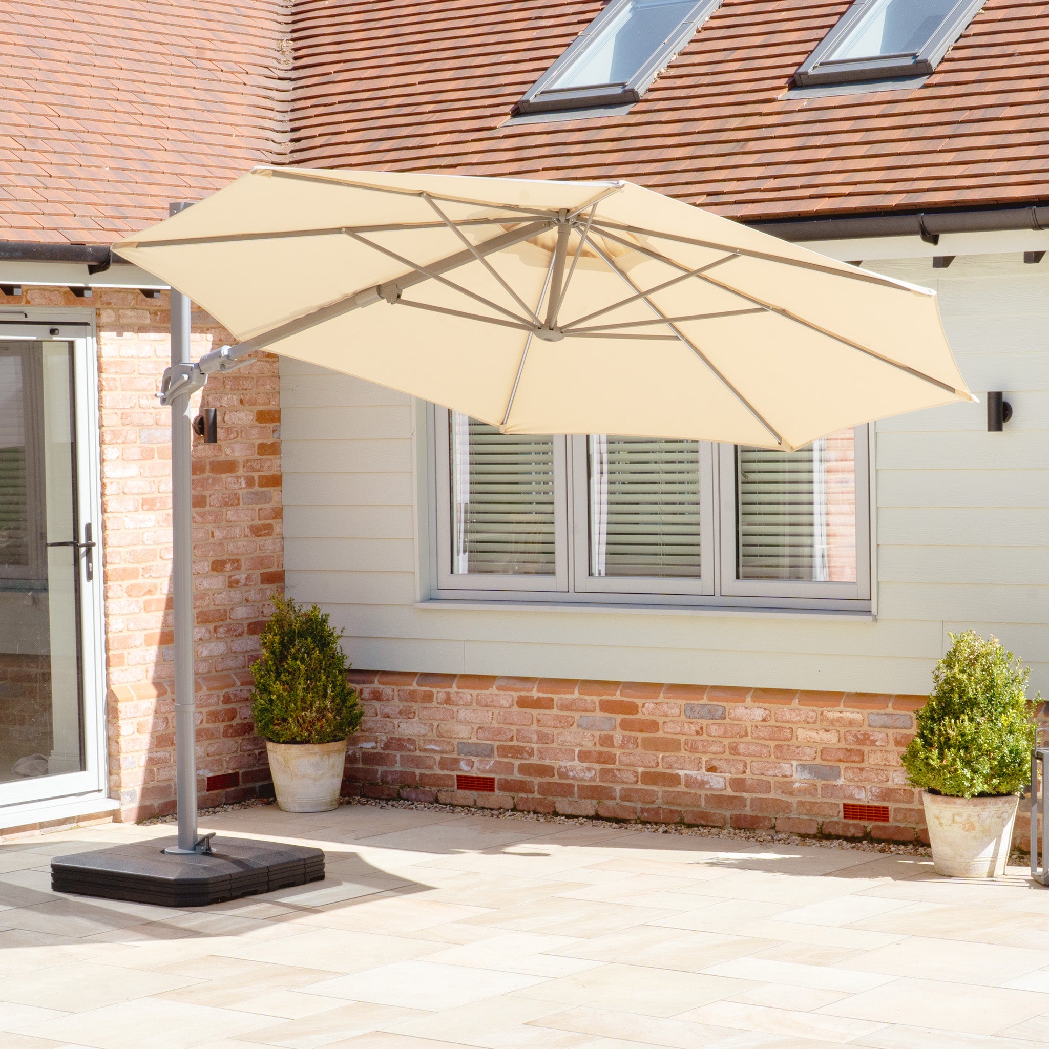 Patio area with a Hera 3m Round Cantilever Parasol in Beige featuring UV50+ fabric and LED lighting, surrounded by brick and siding walls, with potted plants near a window.