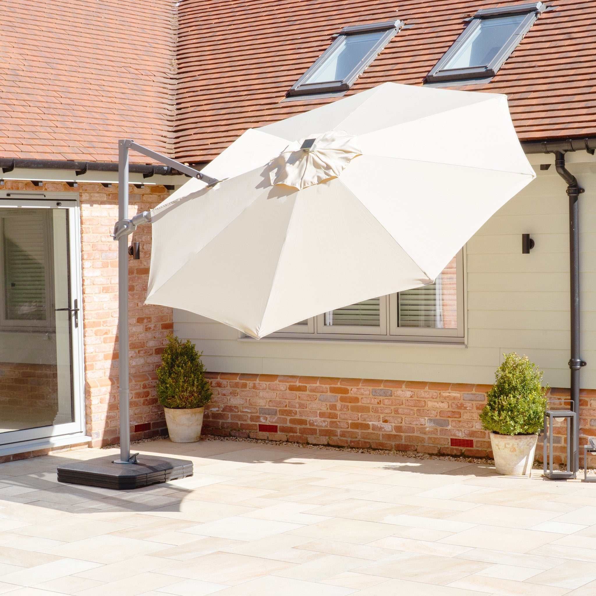 A Hera 3m Round Cantilever Parasol with LED Lighting, in beige, shades a tiled terrace outside a house with brick and light siding, offering UV50+ fabric protection.