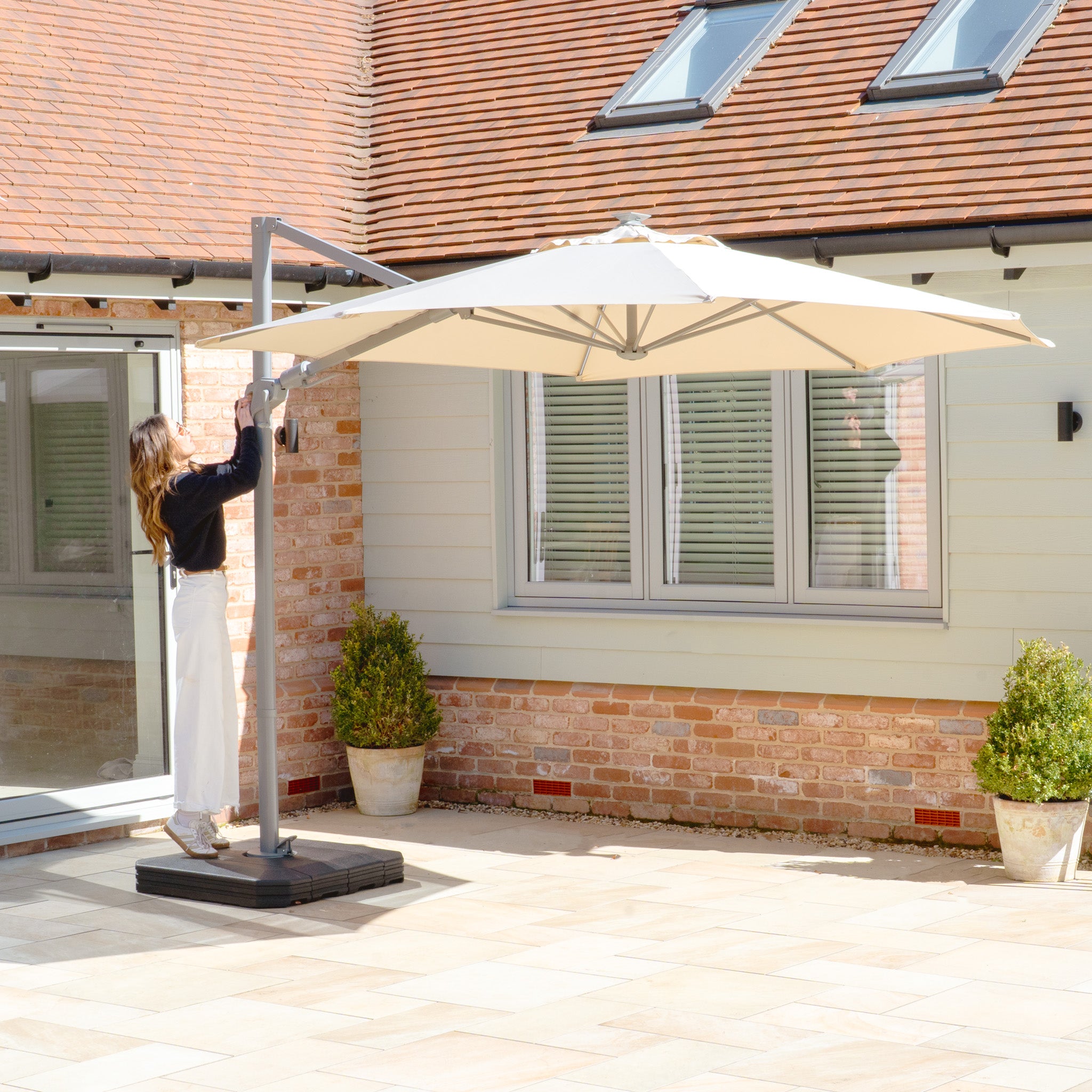 A woman adjusts a Hera 3m Round Cantilever Parasol with LED Lighting in Beige on a patio next to a brick house with a tiled roof.