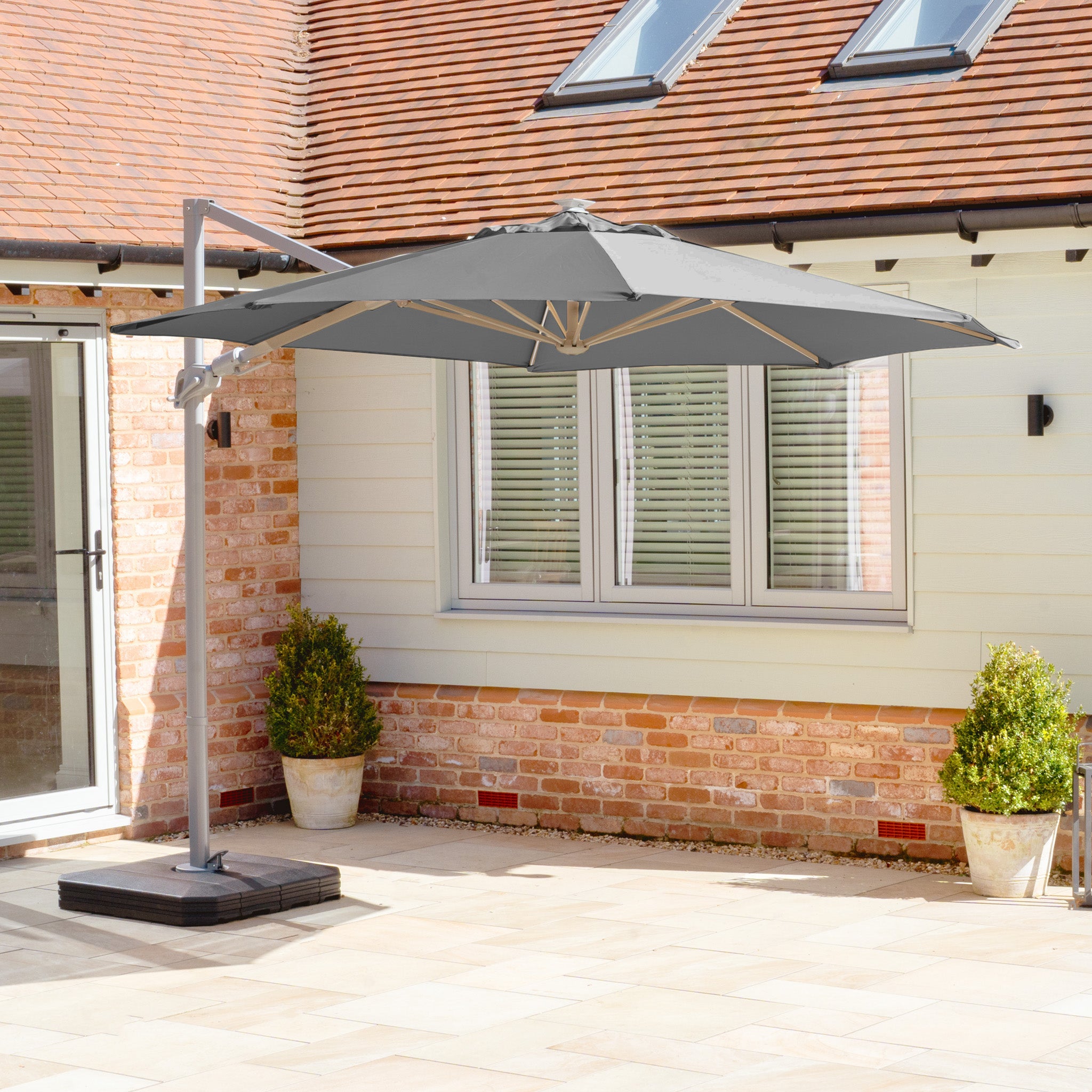 A tranquil patio with the Hera 3m Round Cantilever Parasol in Charcoal, equipped with LED lighting and UV50+ resistance, compliments a charming brick-and-siding exterior. Nearby potted plants enhance the natural elegance of this serene space.