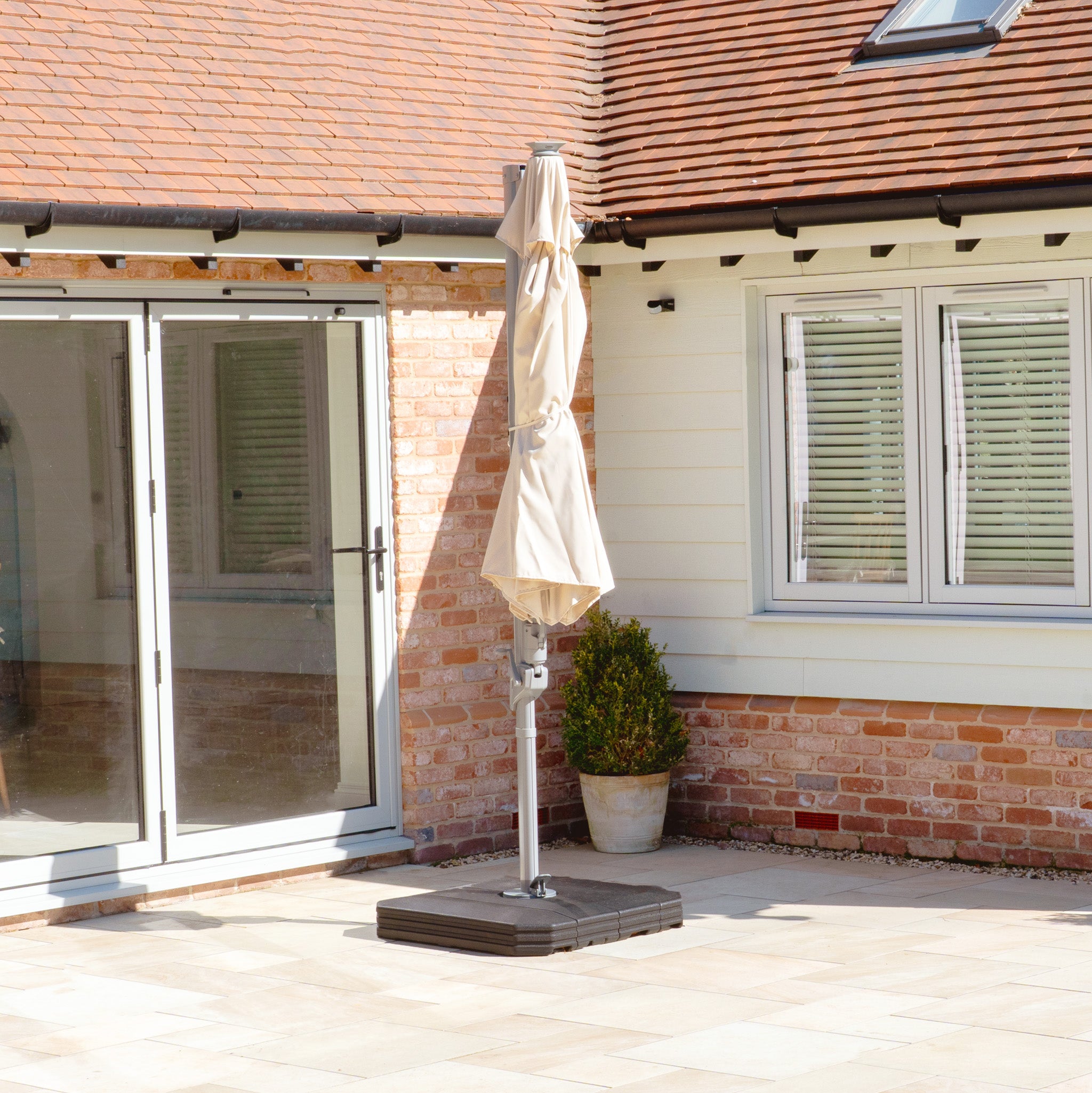 A charming patio displays the Hera 3m Round Cantilever Parasol with integrated LED lighting in Beige, paired with sleek glass doors against a rustic brick wall. Ambient LED lights enhance the setting, complemented by a small plant in front of the window.