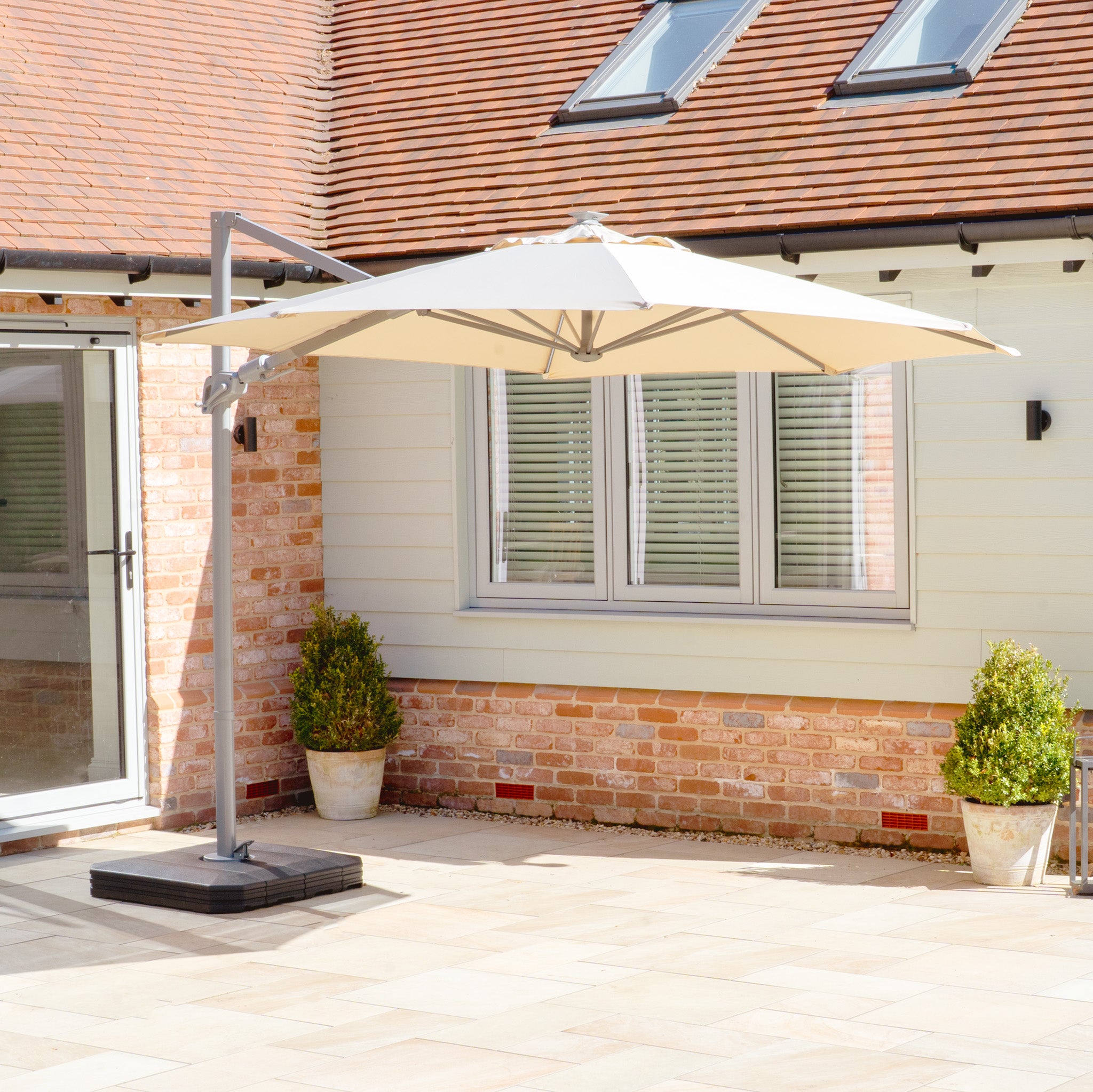 Patio featuring the Hera 3m Round Beige Cantilever Parasol with LED Lighting for UV50+ protection, surrounded by brick and cream walls, with windows and two potted plants near a glass door.