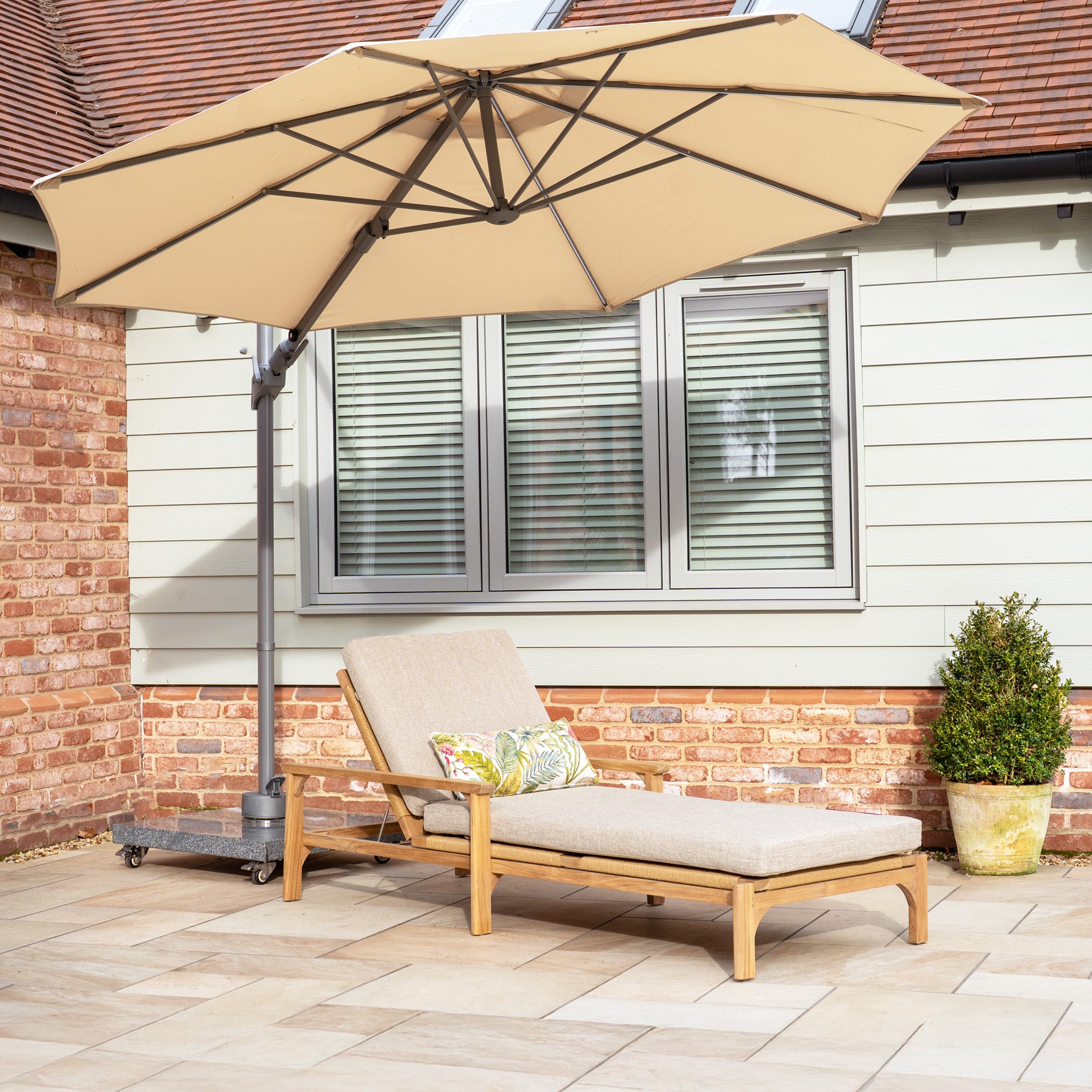 A lounge chair with cushions sits under the beige Hera 3m Round Cantilever Parasol with LED lighting, offering UV50+ resistance and providing shade on a sunny patio by a brick and panel-sided house.