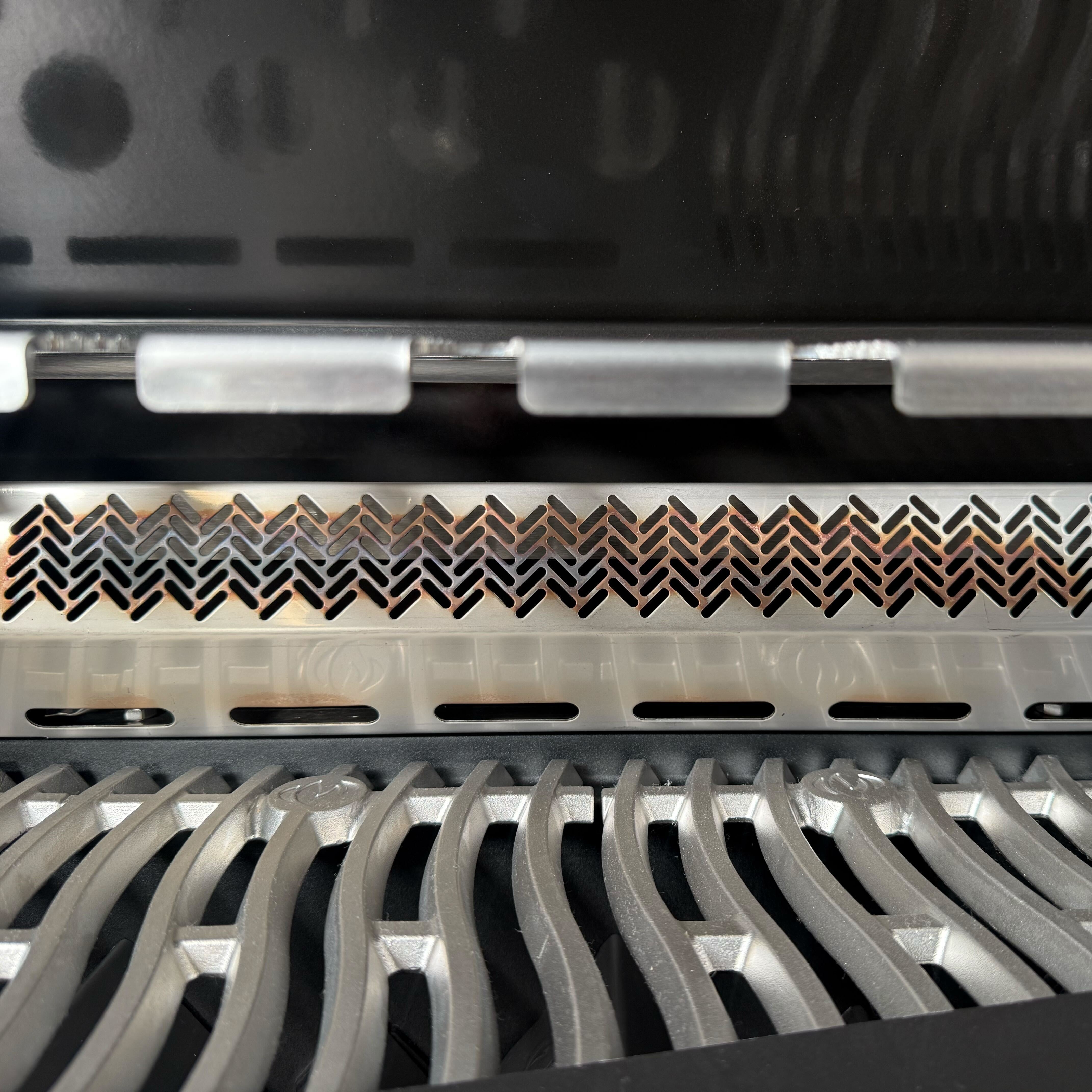 Close-up of a metallic grill or vent with intricate patterns and wavy silver bars below, reminiscent of the stainless steel cooking grids on the Napoleon Phantom Prestige 500 RSIB Gas Barbecue (Ex Display).