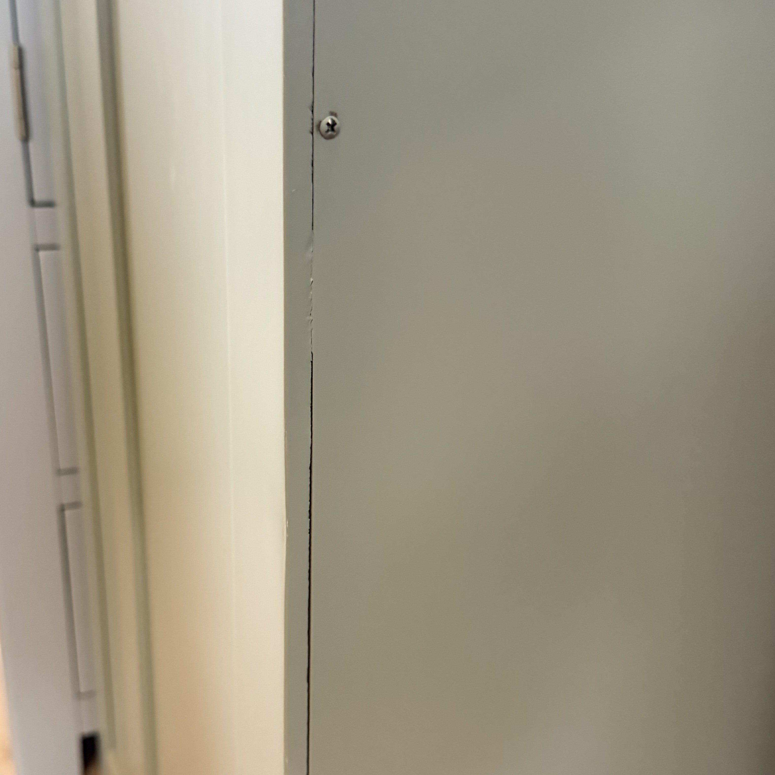 Close-up of a gray metal panel fixed to a wall with a screw near the top, next to the Burford Triple Larder Unit in Sage Green (Ex Display), ideal for kitchen storage.