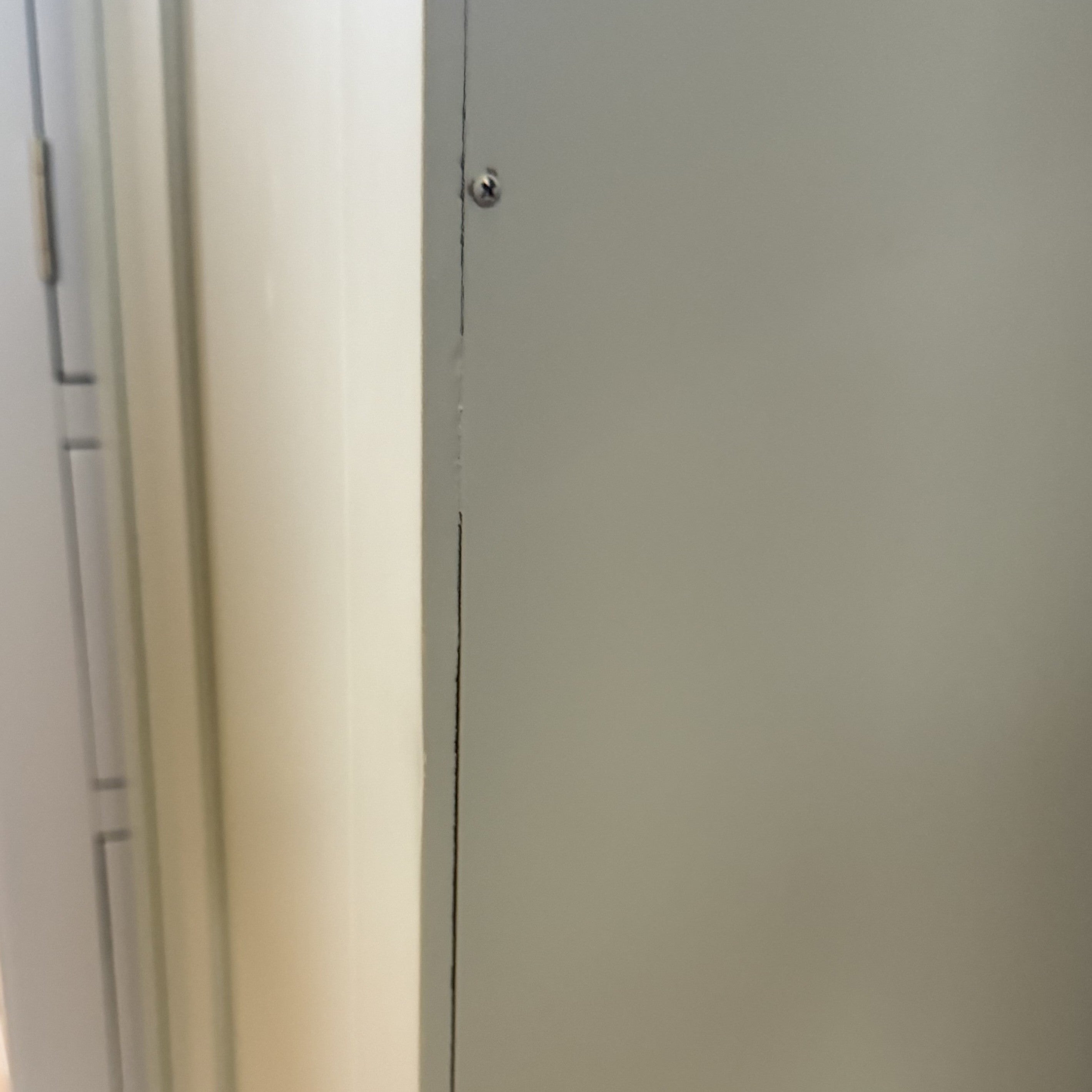 Close-up of a gray metal panel beside a white wall with a visible screw—part of kitchen storage next to the Burford Triple Larder Unit in Sage Green (Ex Display).