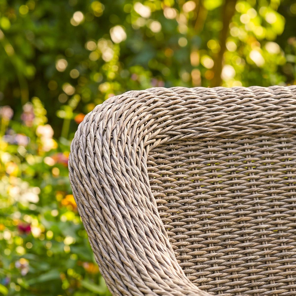 Close-up of the Isla Corner Sofa Set with Rising Table in Flint, highlighting the elegant craftsmanship of its curved wicker backrest, with a blurred garden in the background.