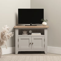 A small TV sits on the Burford Corner Media Unit in Pebble Grey with plants and decor, against a light wall, blending modern style with subtle rustic charm.