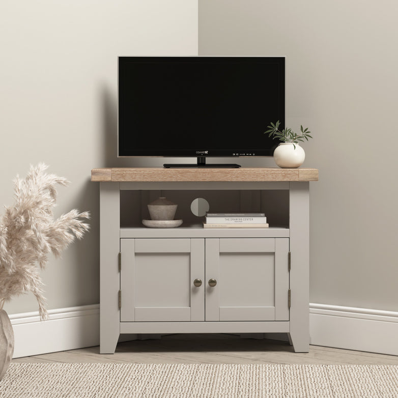 A small TV sits on the Burford Corner Media Unit in Pebble Grey with plants and decor, against a light wall, blending modern style with subtle rustic charm.