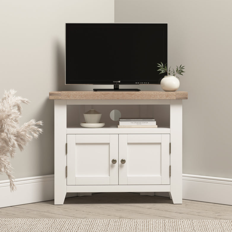 The Burford Corner Media Unit in Warm White holds a small TV, plant, bowl, and books on its light wood and white cabinet. With cable management and ample storage, it keeps your neutral room tidy and organized.