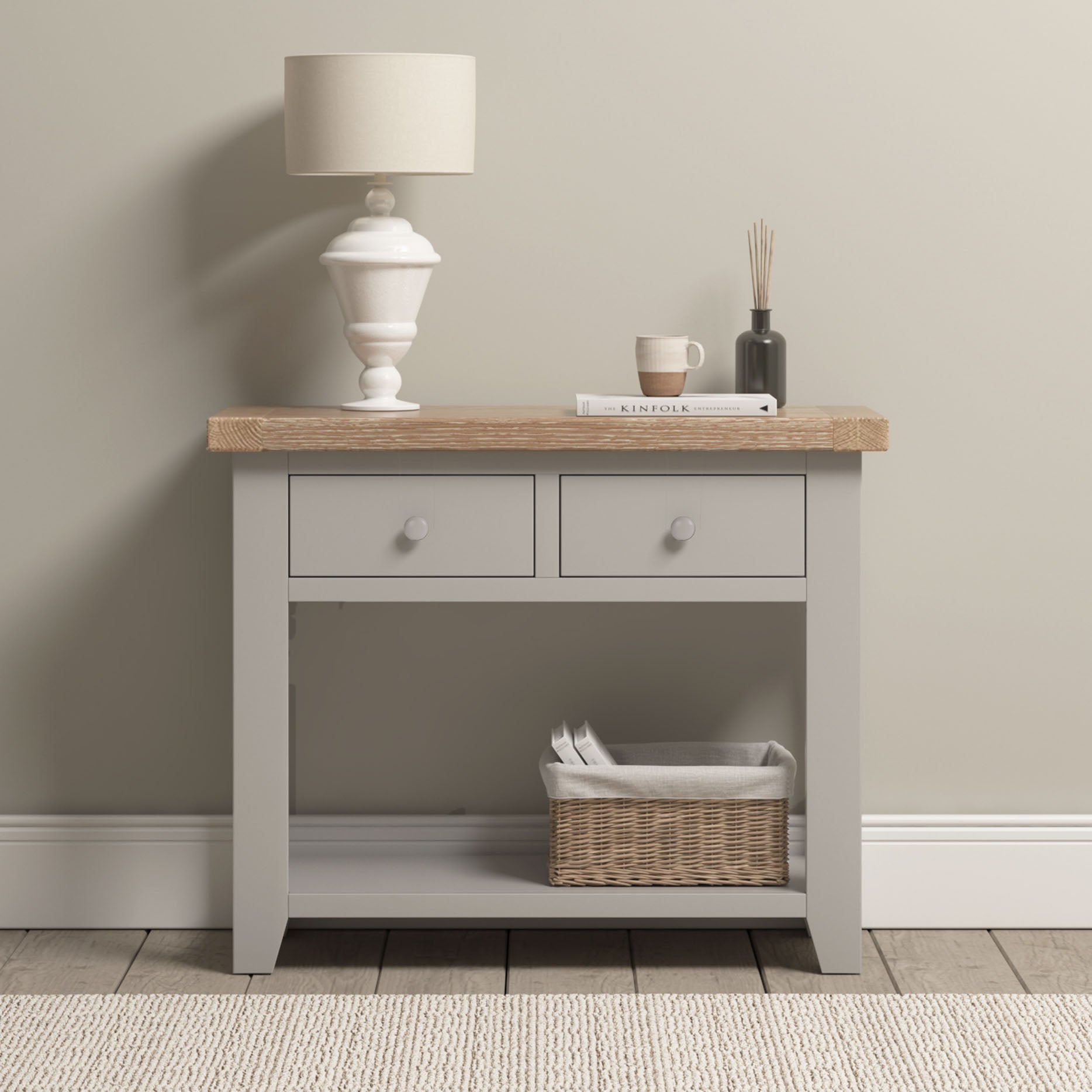 A Burford 2 Drawers Console Table in Pebble Grey with a lamp, books, a cup, and a wicker basket on its natural oak lower shelf.