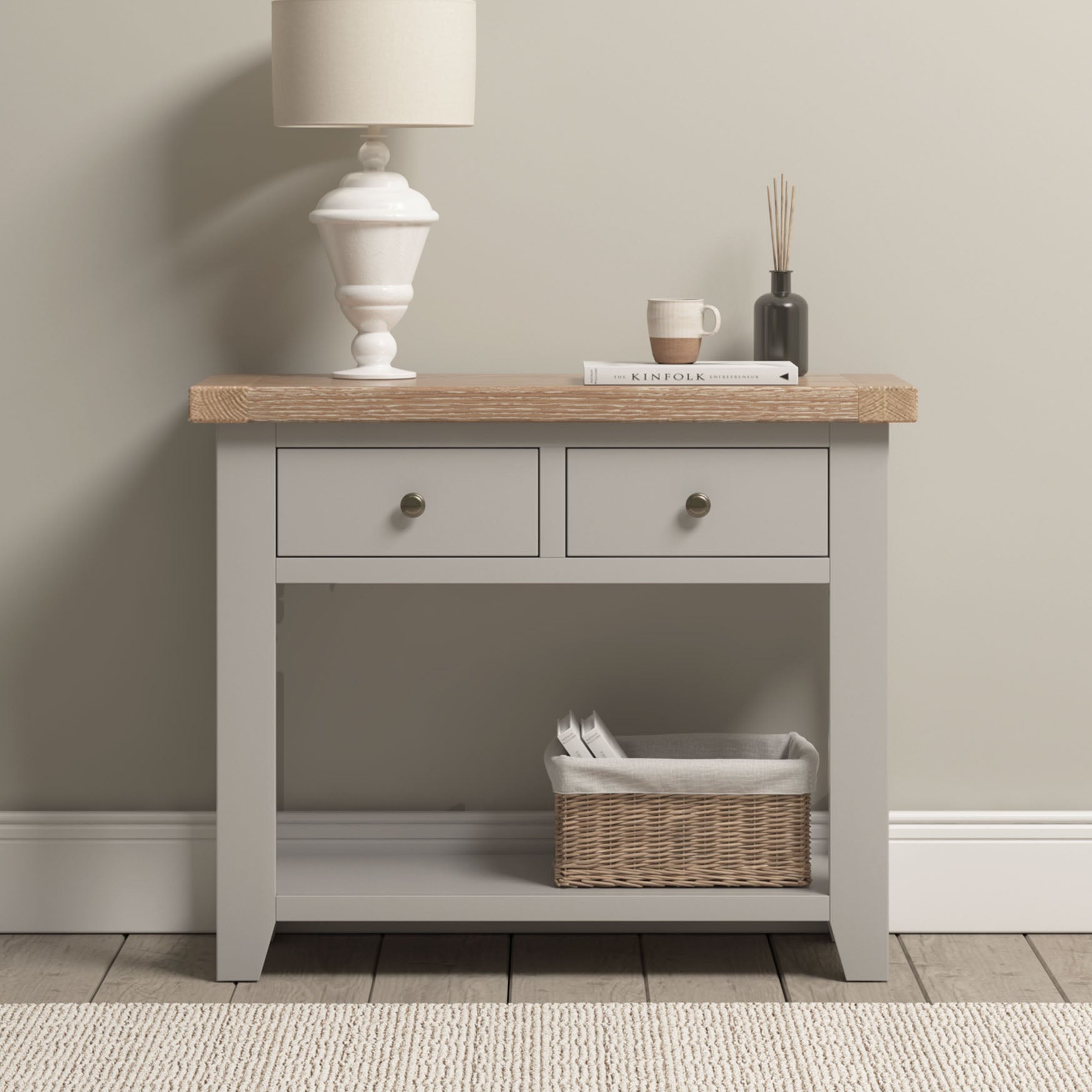 A Burford 2 Drawers Console Table in Pebble Grey with a lamp, cup, book, diffuser on top and a basket on the lower shelf.
