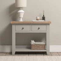 A Burford 2 Drawers Console Table in Pebble Grey with a lamp, cup, book, diffuser on top and a basket on the lower shelf.